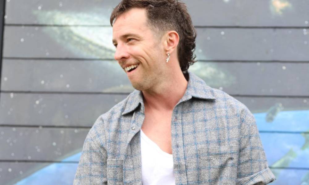 Eli Matthewson stares off camera camera while wearing a grey plaid-patterned shirt layered on top of a white sleeveless shirt. He is standing outside in front of a building with grey and blue designs on it