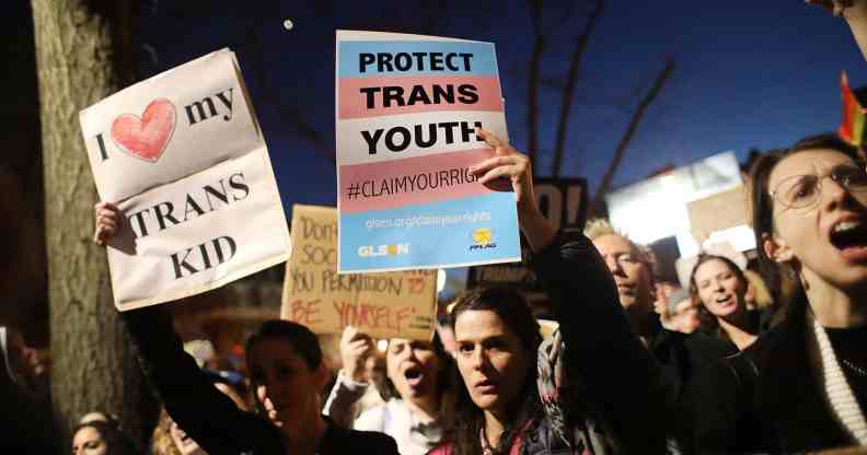 A person is in a gathering amid a crowd in a protest holding a sign that reads 'Protect trans kids'