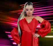 In this photograph, Drag Race Philippines host Jiggly Caliente wears a red sleeveless dress with matching red fabric pieces on her forearms. She has long blonde and brown hair styled in a ponytail and is posed with one hand on her hip. She is standing in front of a dark background with lines of pink, red and yellow