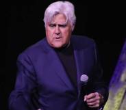 Jay Leno holds a microphone in his hand as he wears a black shirt and dark blue suit jacket as he stands on stage