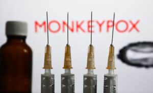 Medical syringes and a bottle are seen with 'Monkeypox' sign and monkeypox illustrative model displayed on a screen in the background in this illustration photo taken in Krakow, Poland.