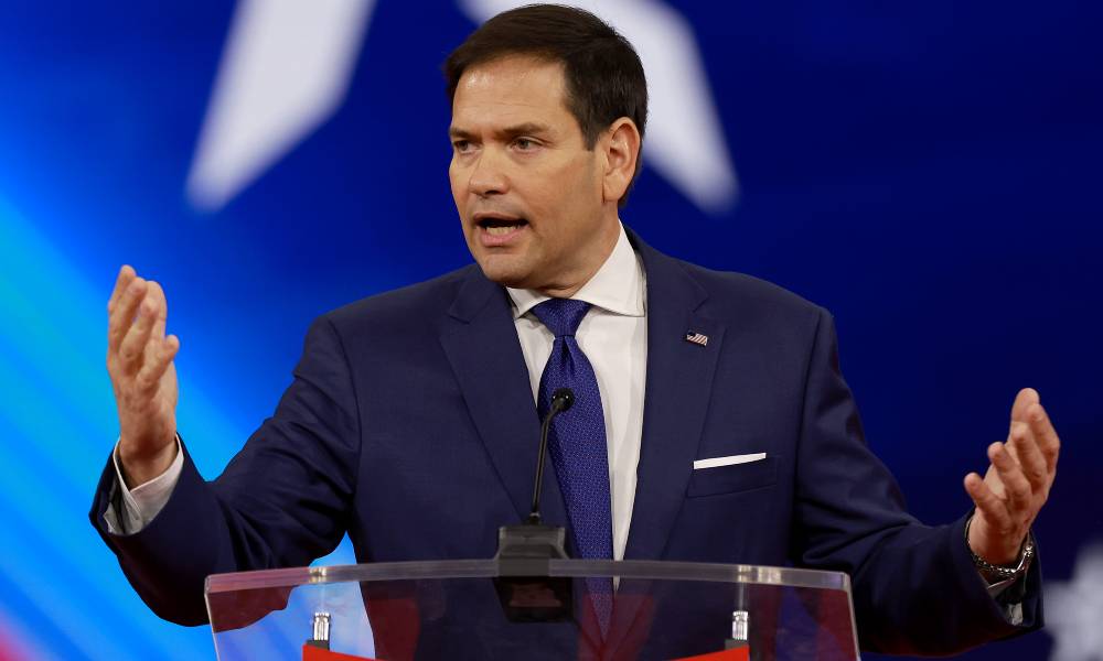 Marco Rubio wears a white shirt, dark blue tie and dark suit jacket as he stands at a podium. He is gesturing with both hands as he speaks to a crowd off camera