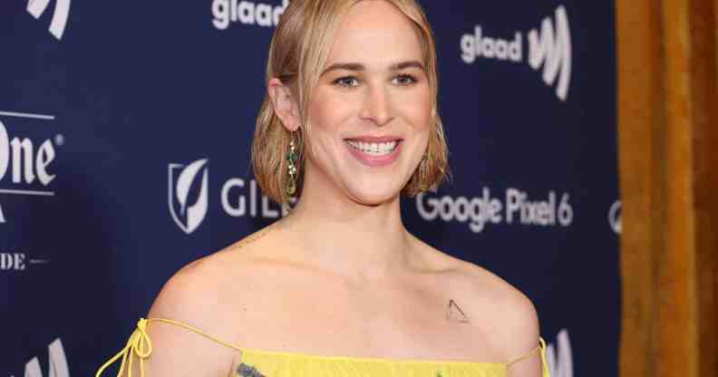 Tommy Dorfman stands in the press section of the GLAAD Media Awards wearing a yellow dress.