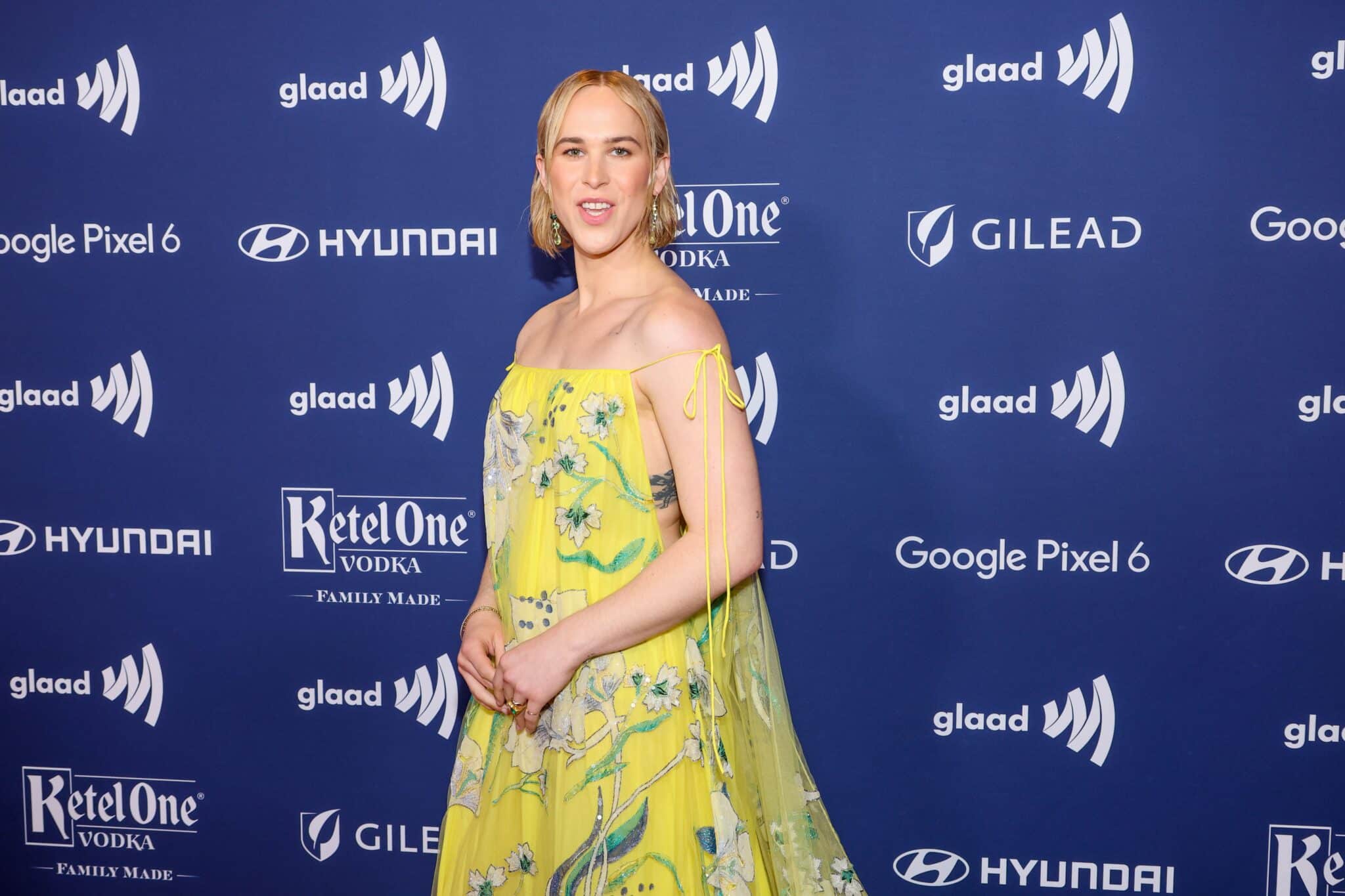 Tommy Dorfman posing behind a blue GLAAD Media screen in a yellow shoulderless dress.