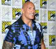 Dwayne 'The Rock' Johnson smiles for the camera as he wears a blue and black patterned top and stands in front of a patterned background with the SD Comic Con logo on it