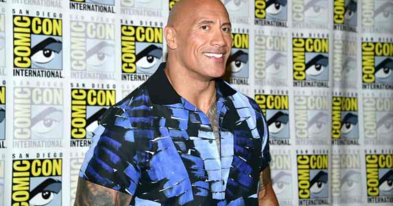 Dwayne 'The Rock' Johnson smiles for the camera as he wears a blue and black patterned top and stands in front of a patterned background with the SD Comic Con logo on it