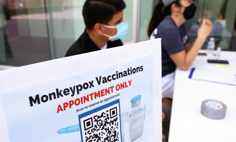 A sign is seen outside with the words 'Monkeypox vaccinations appointment only' on it with a QR code beneath. There are people sitting behind a table behind the sign