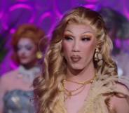 Drag Race Down Under season two queen Faúx Fúr looks off camera while wearing a cream outfit with other queens standing in the background