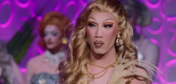 Drag Race Down Under season two queen Faúx Fúr looks off camera while wearing a cream outfit with other queens standing in the background