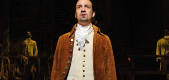 Lin-Manuel Miranda wears a white shirt, cream coloured waist jacket and brown coat as he performs in Hamilton