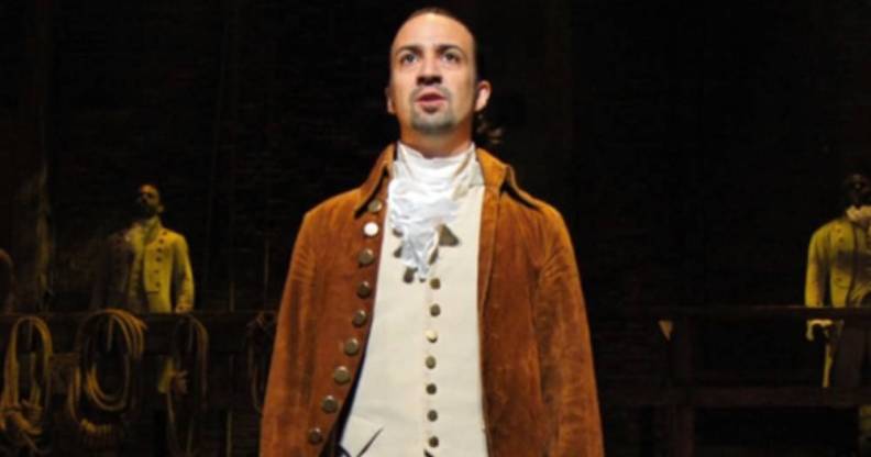 Lin-Manuel Miranda wears a white shirt, cream coloured waist jacket and brown coat as he performs in Hamilton