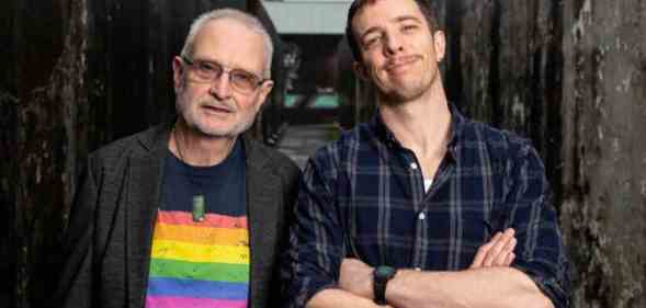 Eli Matthewson wears a dark, plaid patterned shirt as he crosses his arms in front of his chest. He is standing next to his dad who is wearing a shirt with a rainbow LGBTQ+ pride flag on it