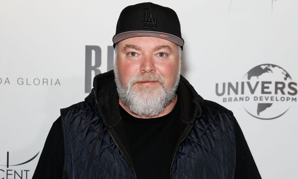 Kyle Sandiland wears a dark outfit and hat as he stands in front of white background