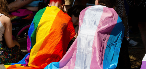 Two people sit side by side while wearing an LGBTQ+ and trans pride flag around their shoulders