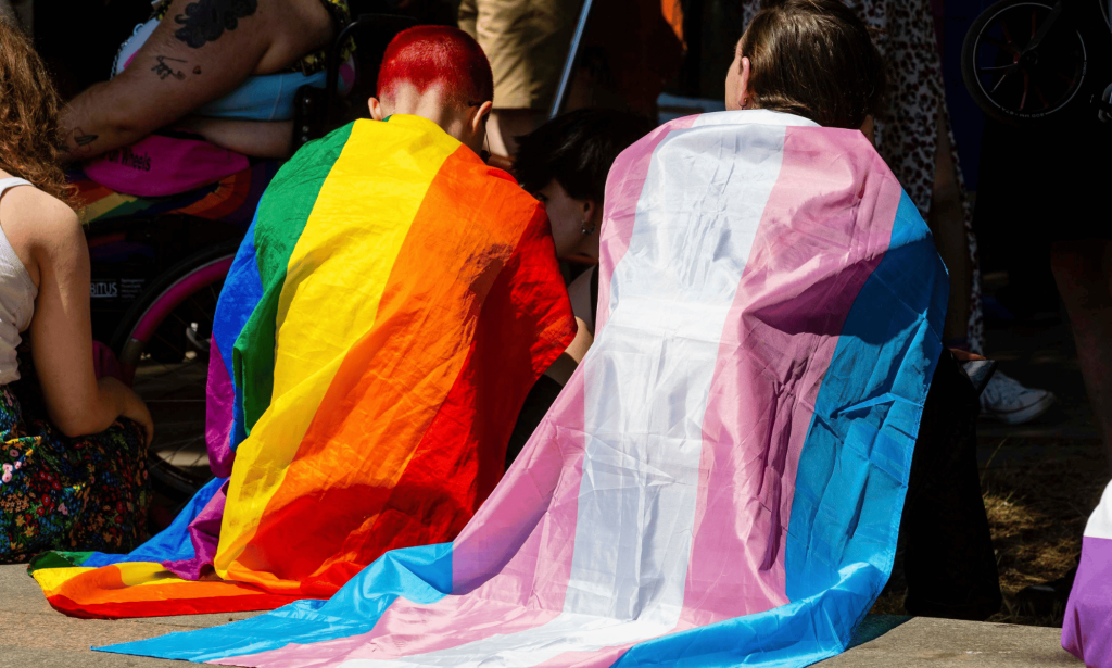 Two people sit side by side while wearing an LGBTQ+ and trans pride flag around their shoulders