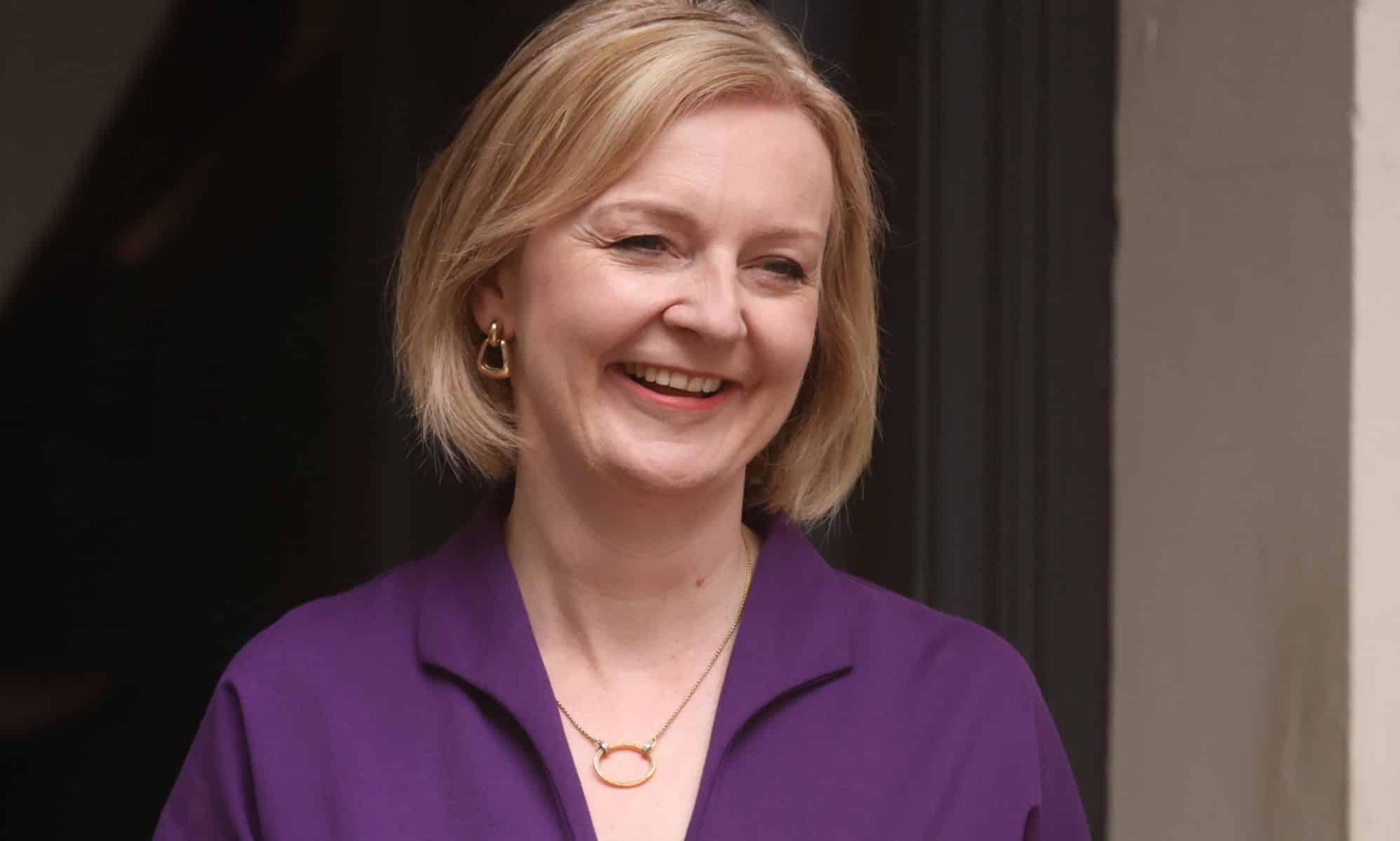 Liz Truss smiles as she stands outside a building wearing a purple outfit