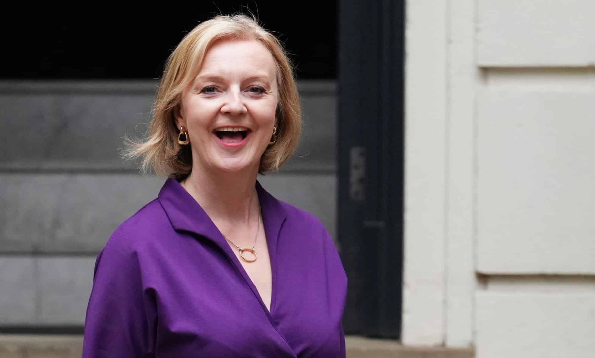 Liz Truss smiles as she stands outside a building wearing a purple outfit