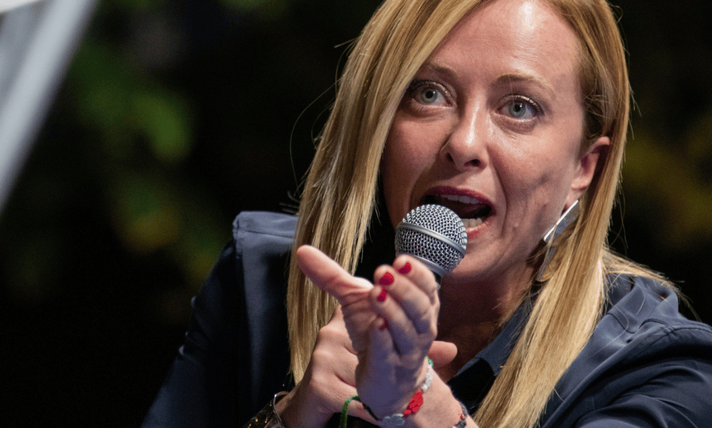 Giorgia Meloni wears a dark outfit as she speaks into a microphone and gestures with her hand