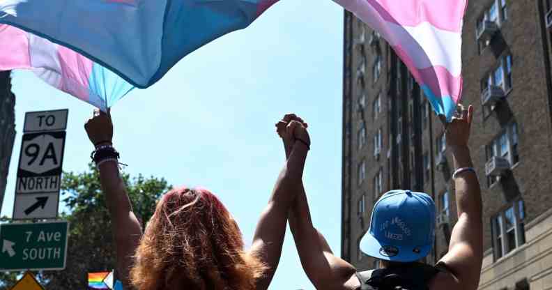 Two trans people hold hands while marching under a trans pride flag.