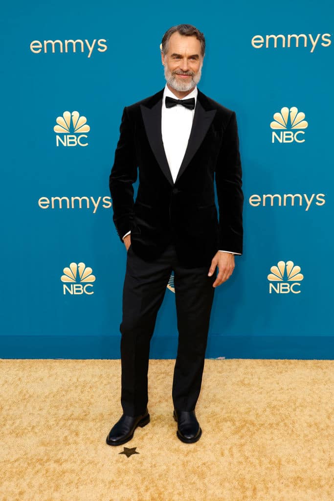 Murray in a black suit and white shirt, stood against a blue wall on a gold carpet