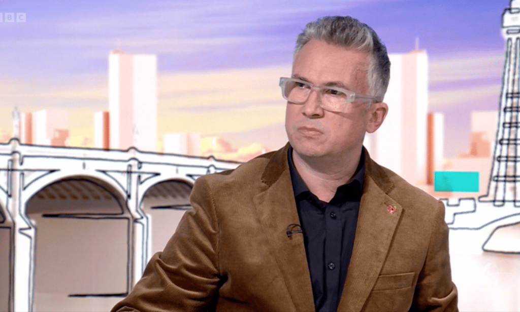 Iain Anderson wears a black shirt and brown jacket as he does an interview with Laura Kuenssberg on the BBC