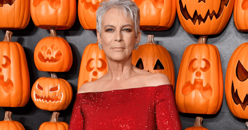 Jamie Lee Curtis wears a red outfit as she stands in front of a wall filled with orange pumpkin items shaped to look like jack-o-lanterns