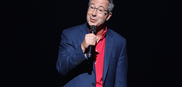 Ben Elton wears a suit while speaking into a microphone on stage