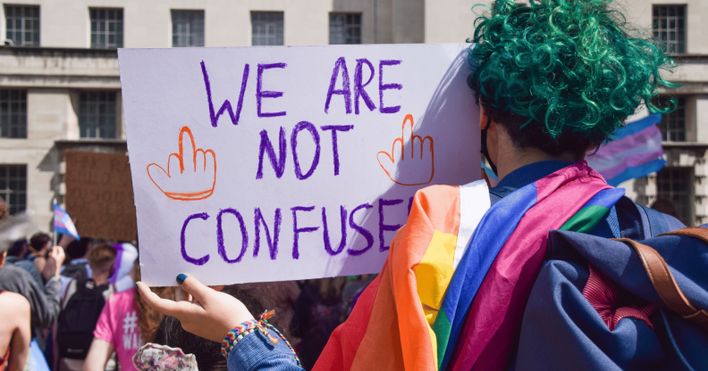 A person with green hair, who has their back turned to the camera, holds up a sign reading 'we are not confused' with an LGBTQ+ Pride flag draped along their shoulders