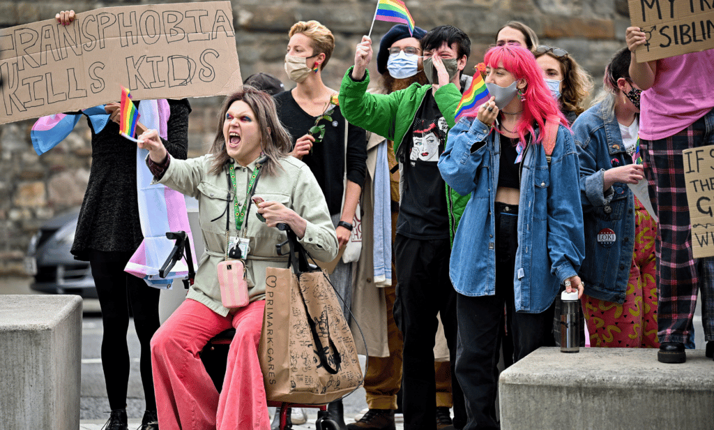 Several people gather in a crowd to protest in support of the trans community, one person holds up a sign reading 'transphobia kills kids'