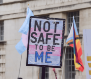 A person holds up a sign, designed in the colours of the trans Pride flag, that reads 'not safe to be me'