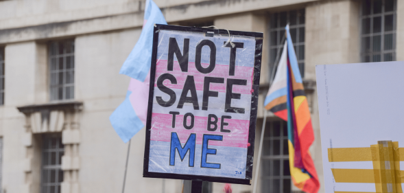 A person holds up a sign, designed in the colours of the trans Pride flag, that reads 'not safe to be me'