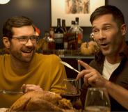 Billy Eichner and Luke Macfarlane at a dinner table, laughing with cutlery raised mid-gesture, in Bros