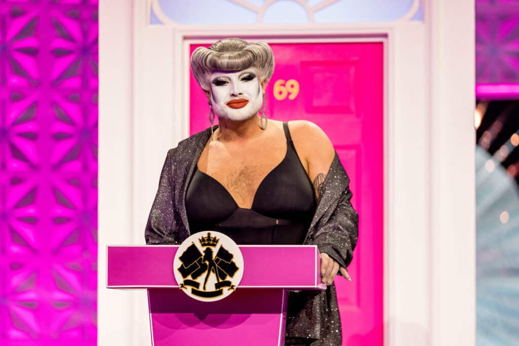 Drag Race UK queen Danny Beard standing at a pink podium in front of a pink front door