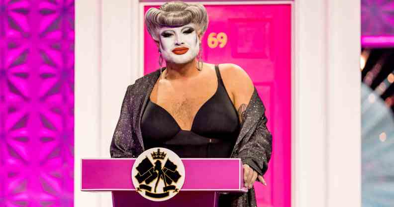 Drag Race UK queen Danny Beard standing at a pink podium in front of a pink front door