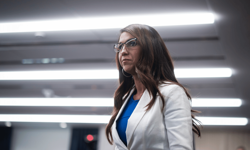 Colorado representative Lauren Boebert wears a white jacket and blue outfit as she stands in a room