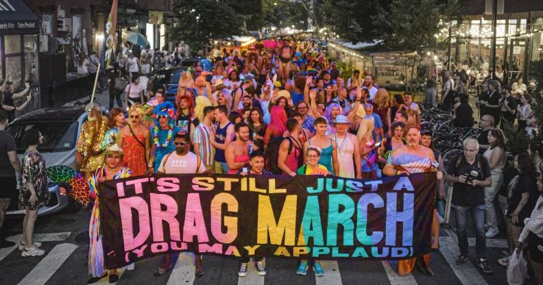 A photo of pro-drag march with people at the front of the crowd holding a banner with the slogan "It's still just a drag march, you may applaud".