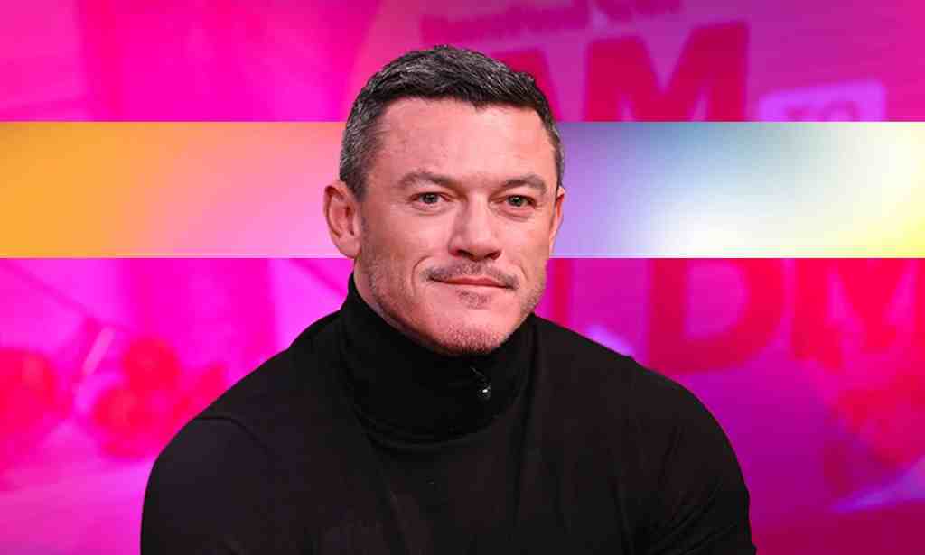 Actor Luke Evans wearing a black top sits in front of a pink background