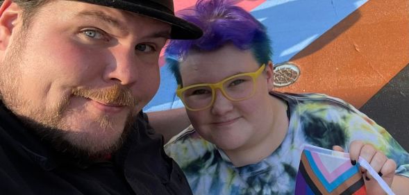 A photo of chef Dave Heide dressed in black and wearing a black hat with his arm round his child Ollie who's wearing a tie-dye t-shirt, yellow glasses and is holding a Pride flag