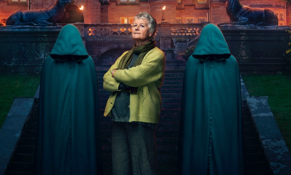 A promo photo from BBC's The Traitors showing contestant Andrea dressed in a dark green top and light green cardigan with her arms crossed standing on some steps to a gothic-looking building with two people standing either side of her wearing hooded cloaks and their heads bowed so as to hide their faces. (BBC/Studio Lambert Associates/Mark Mainz)