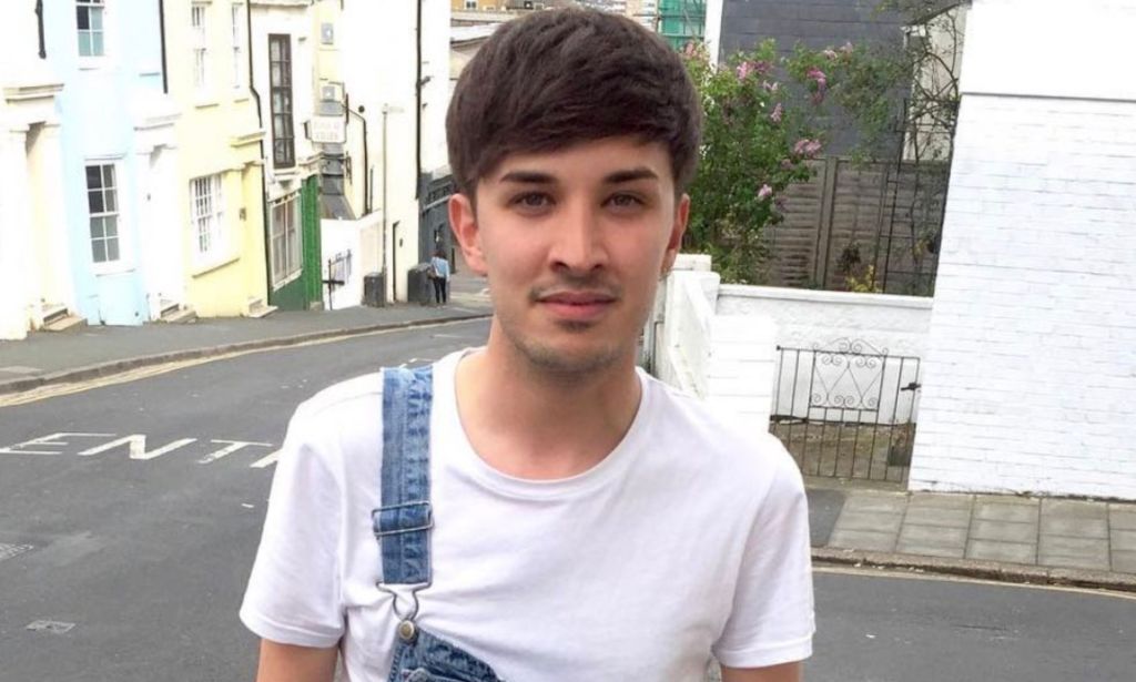 A photo of one of the Manchester Arena bombing victims, Martyn Hett, wearing a white t-shirt and dungerees with one strap, smiling outside a street corner.