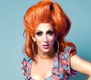 Marti G. Cummings wears a red wig while in drag, and a dress with slightly worn American flags across it, standing in front of a blue background.