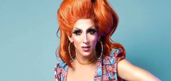 Marti G. Cummings wears a red wig while in drag, and a dress with slightly worn American flags across it, standing in front of a blue background.