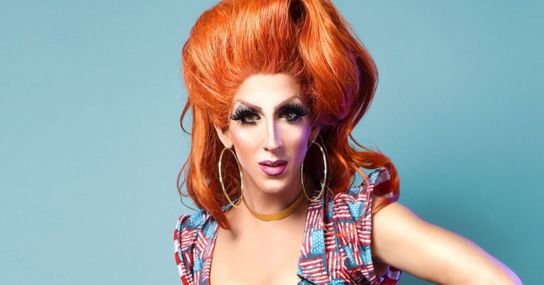 Marti G. Cummings wears a red wig while in drag, and a dress with slightly worn American flags across it, standing in front of a blue background.