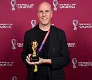 Grant Wahl stands infront of a red back drop with the Qatar World Cup logo written across it, while holding a miniature replica of the World Cup trophy, wearing a black blazer.