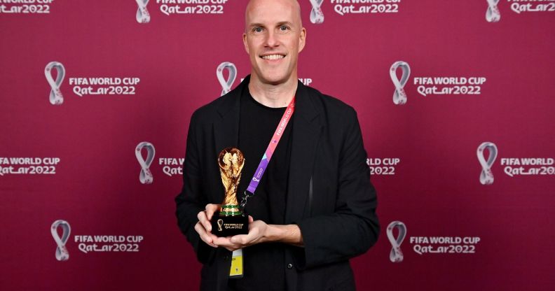 Grant Wahl stands infront of a red back drop with the Qatar World Cup logo written across it, while holding a miniature replica of the World Cup trophy, wearing a black blazer.