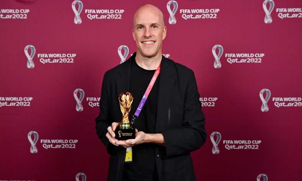 Grant Wahl stands infront of a red back drop with the Qatar World Cup logo written across it, while holding a miniature replica of the World Cup trophy, wearing a black blazer.