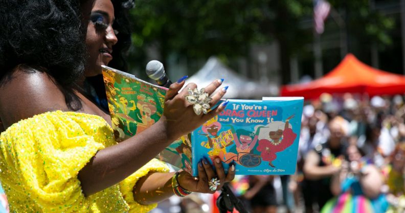 A drag queen, wearing a yellow dress, reads to a crowd from a book titled "if you're a drag queen and you know it."