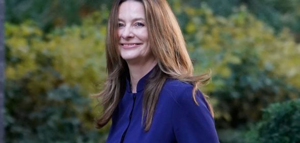 Gilian Keegan, in a navy blue dress, walks towards number 10 downing street by nearby foliage, which is blurred in the background.