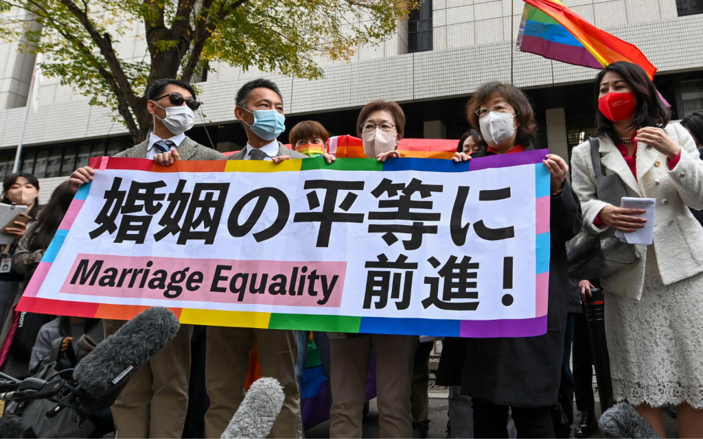 Plaintiffs holding up a rainbow-themed banner supporting marriage equality as a rainbow flag waves in the background.