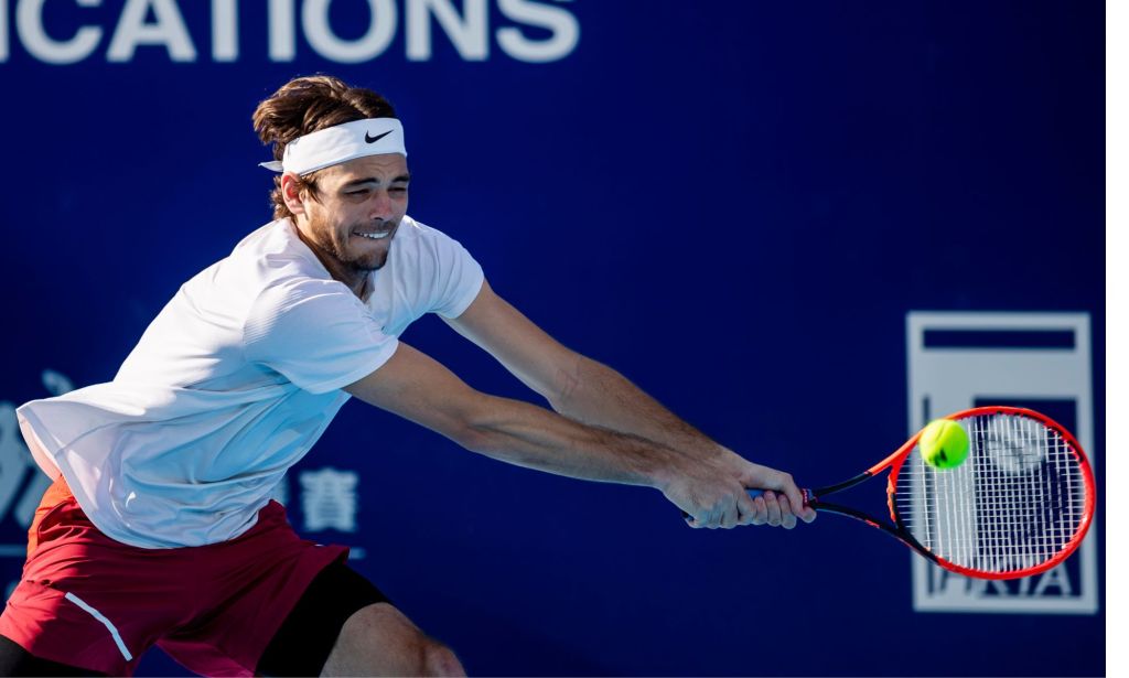 Taylor Fritz, wearing a white top, lunges for a tennis ball with a red racket in his hand while playing an ATP match.